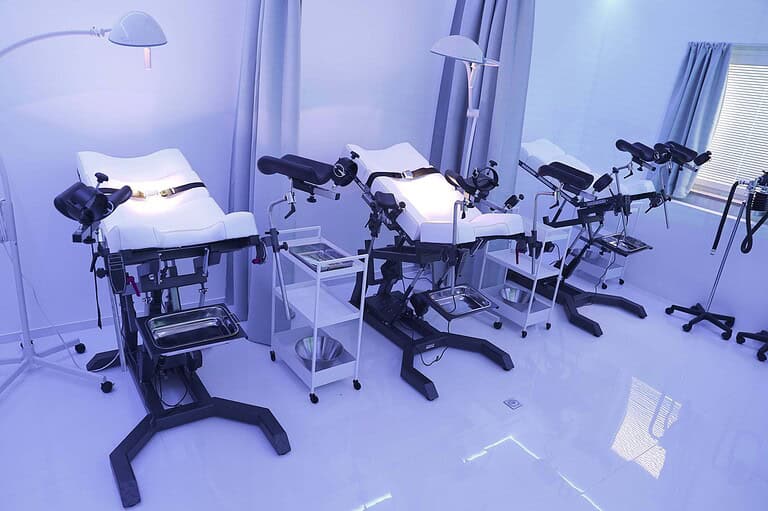 Helle, moderne BDSM-Medizinzimmer mit Liegen, Lampen und medizinischen Geräten.